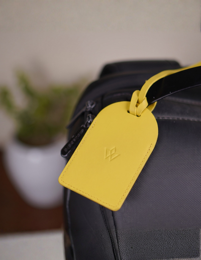 A bright yellow leather luggage tag with an embossed LV logo, attached to a black bag strap, shown in a close-up shot with a blurred background.