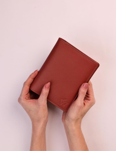 A person's hands holding a reddish-brown leather wallet against a solid pale pink background.