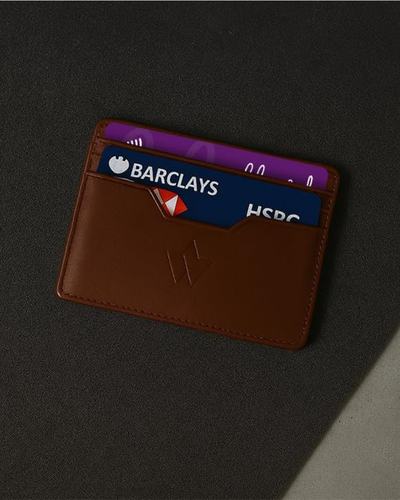 A brown leather cardholder wallet with three cards partially inserted, photographed on a dark textured surface with a diagonal light shadow.