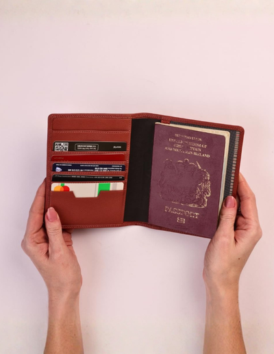 A person's hands holding open a reddish-brown leather passport holder containing credit cards and a US passport, against a plain light pink background.