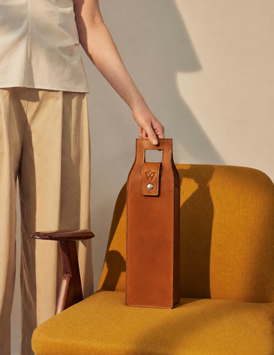 A person in a light beige outfit holds the handle of a brown leather wine bag standing on a mustard yellow armchair against a warm beige wall.