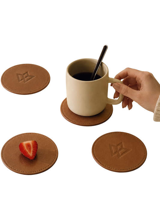 Four round brown leather coaster mats with a debossed geometric logo, shown on a white background. One coaster is under a beige ceramic mug with a spoon, and another coaster holds a strawberry slice.