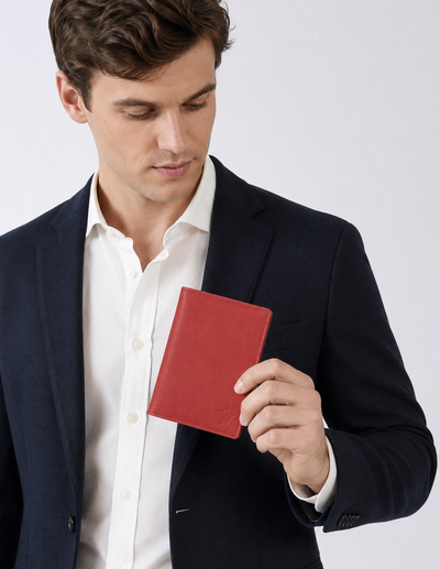 A man in a navy blazer and white dress shirt holds a small red leather passport cover or notebook against a light gray studio background.