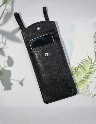Black pebbled leather phone pouch with a top loop strap and a front slip pocket holding a smartphone, styled on a white surface with leaf shadows and a plant card.