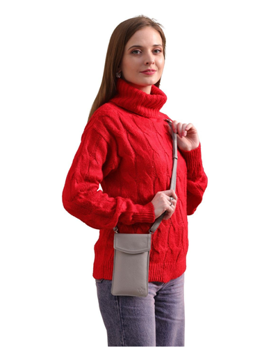 A woman wearing a bright red cable-knit turtleneck sweater and gray jeans, carrying a small taupe crossbody phone bag against a plain white background.