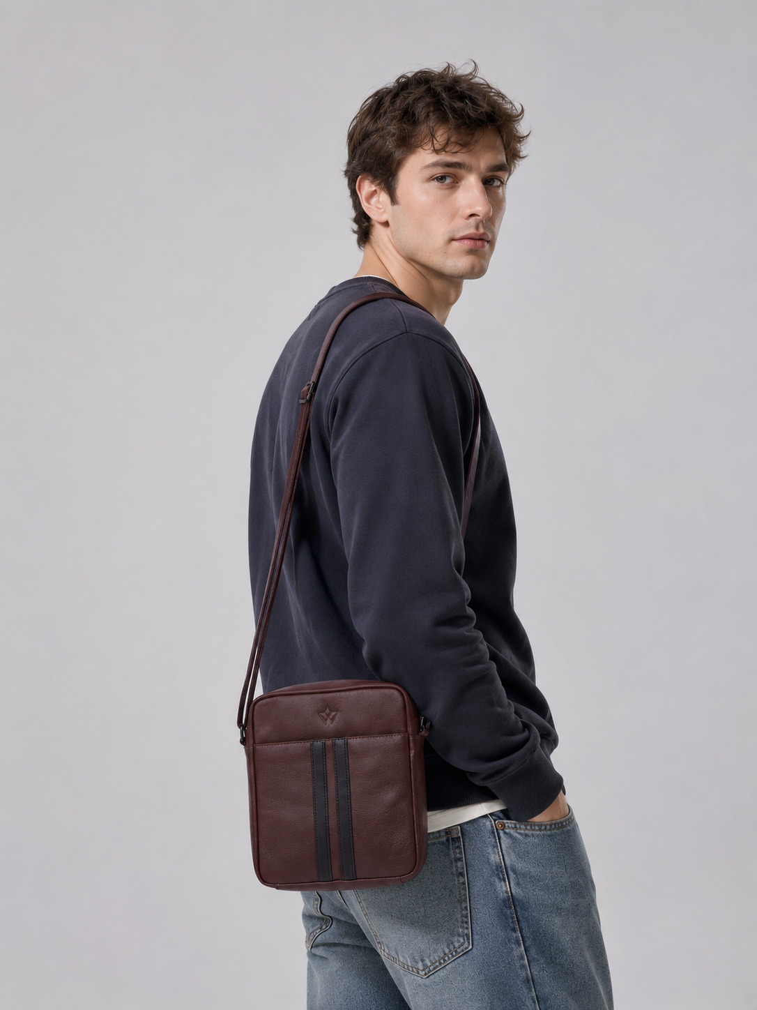 A man wearing a dark navy sweatshirt and blue jeans models a brown leather crossbody bag with two vertical black stripes, shown from a side-back angle against a neutral gray studio background.