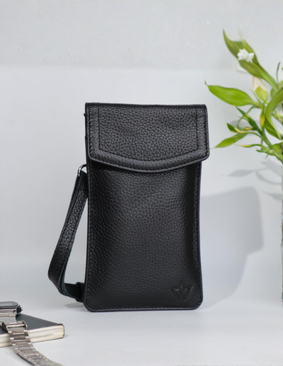 A black pebbled leather crossbody phone pouch with a flap closure, shown standing upright on a light surface with a small plant and books in the background.