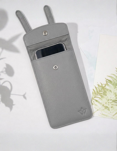A light gray textured leather phone pouch with a top strap and a front pocket holding a smartphone, photographed on a white surface with soft plant shadows and a decorative card at the side.