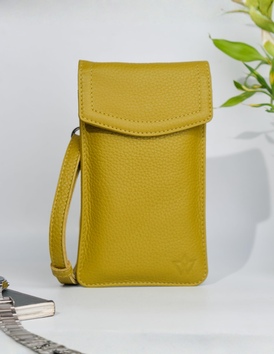 A yellow pebbled leather phone pouch with a flap closure and wrist strap, standing upright on a white surface with a small plant leaf and metallic object in the background.