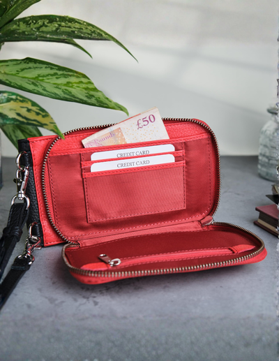 An open red zip-around wallet with multiple card slots, a £50 note, and cash visible, photographed on a gray surface with a plant leaf and decorative objects in the background.