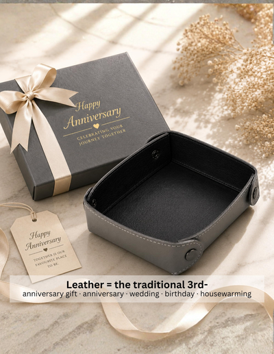 A dark gray leather anniversary gift box shown open next to its matching closed box with a cream ribbon and tag, on a light textured surface with dried flowers and soft window-light shadows.