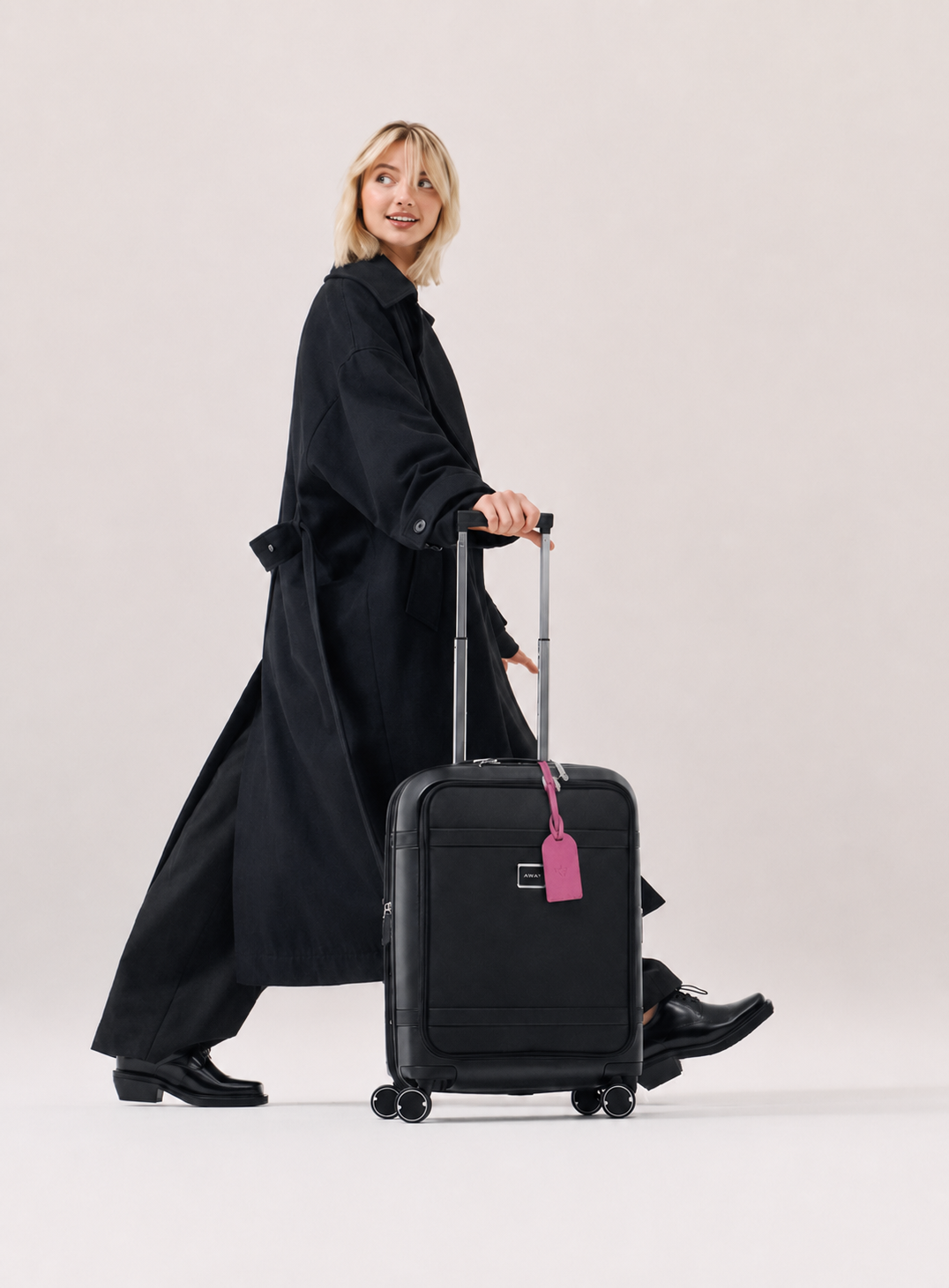 A woman in a long black trench coat pulls a black rolling suitcase with a pink luggage tag against a plain light beige studio background.