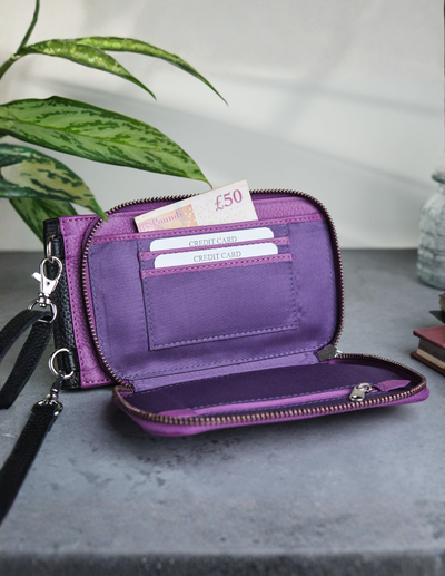 A purple zip-around wallet with an open interior showing card slots, a £50 note, and a wristlet strap, displayed on a gray surface with a plant in the background.