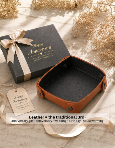 An open brown leather gift box with a dark interior sits beside a black square Anniversary gift box tied with a cream ribbon and a matching paper tag, styled on a light marble surface with dried flowers.