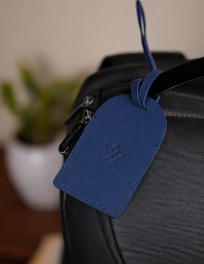 A blue leather luggage tag with an embossed geometric logo, attached to a black backpack strap, with a softly blurred indoor background.