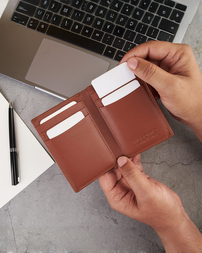 A person's hands holding an open brown leather bifold wallet with card slots and a card inserted, set against a gray desk background with a laptop, pen, and notebook.