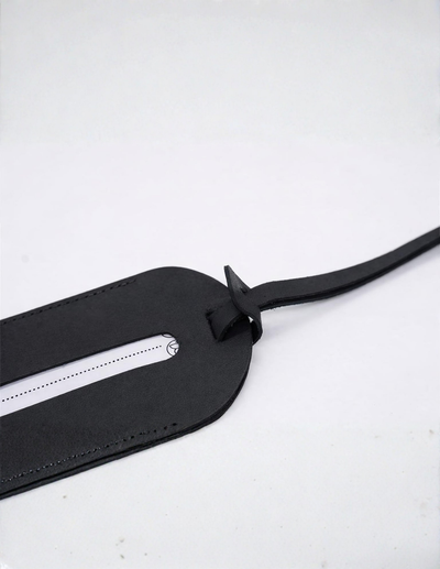 A close-up of a black leather luggage tag with a horizontal slot and a strap, shown on a light gray background.