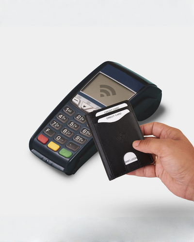 A hand holding a black leather cardholder wallet in front of a black wireless payment terminal on a light gray background.
