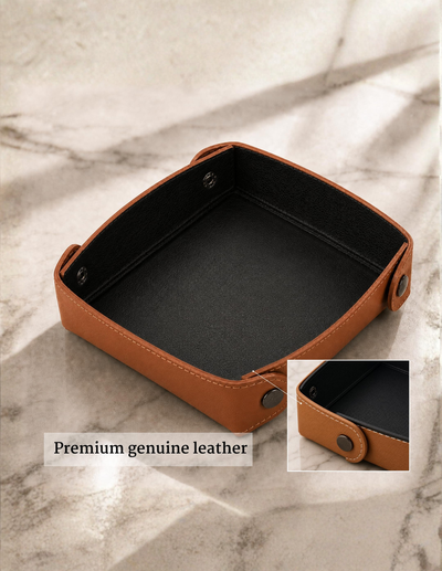 A square tan leather tray or organizer with a black interior, shown on a light marble surface. A small inset at bottom right shows a close-up of the leather edge and snap detail, and text at bottom left reads 'Premium genuine leather'.