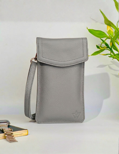 A light gray pebbled leather crossbody phone pouch with a flap closure and wrist strap, standing upright on a white surface with a small embossed logo near the bottom right. A blurred green plant and a small gold-toned accessory are visible in the background.