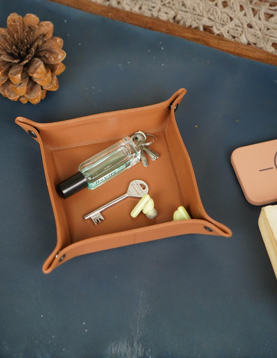 A tan leather square organizer tray holding a perfume bottle, keys, earplugs, and a small keychain, styled on a dark blue surface with a book and pinecone nearby.
