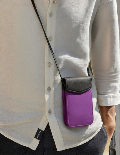 A person wearing a white button-up shirt and dark pants carries a small purple and black crossbody phone pouch with a black strap.