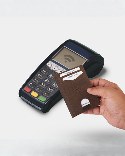 A hand holding a black leather cardholder wallet in front of a brown wireless payment terminal on a light gray background.