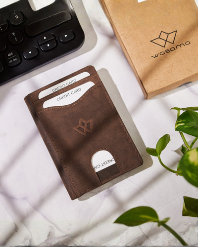 A brown leather card holder wallet with a debossed logo, shown on a white marble surface next to a keyboard, a kraft paper box branded wasamo, and a green plant.