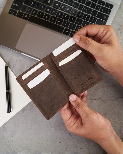 A person holds an open brown leather bifold wallet with card slots, shown over a gray tabletop with a laptop and pen in the background. The wallet has the brand name 'yabomo' embossed on the inside.