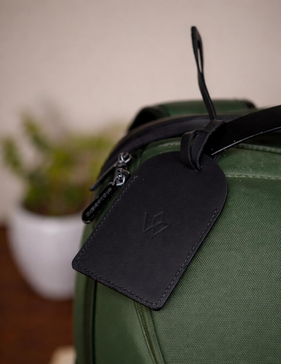A close-up of an olive green canvas bag with a black leather luggage tag attached by a black loop strap. The embossed logo on the tag is partially obscured, but the tag is stitched and shaped like a luggage tag.
