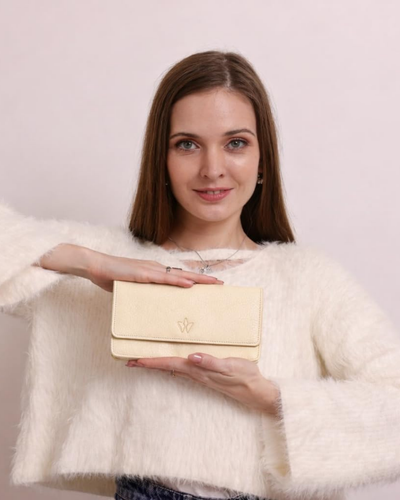 A woman wearing a fluffy cream sweater holds a beige coloured leather clutch bag with a gold monogram logo against a light pink background.