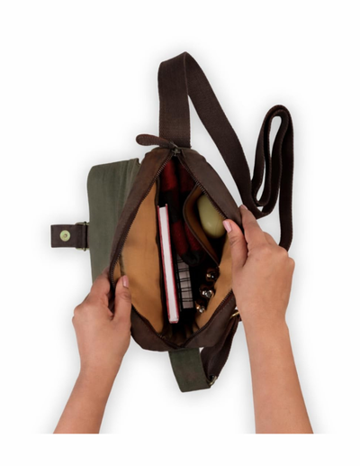 A dark brown crossbody bag with a wide strap, shown open with a person's hands holding the sides to display the interior compartments and contents, set against a plain white background.