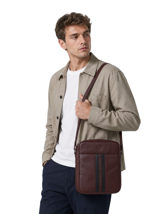 A man wearing a beige overshirt, white t-shirt, and navy trousers carries a dark brown leather crossbody bag with vertical stripes on a plain white background.