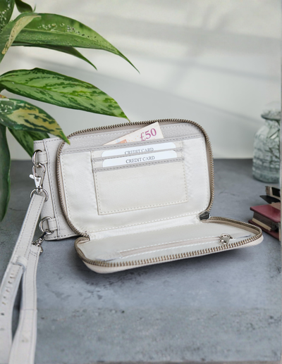 An open cream-colored leather wallet with a wristlet strap, displayed on a gray stone surface with green leaves and a small vase in the background.