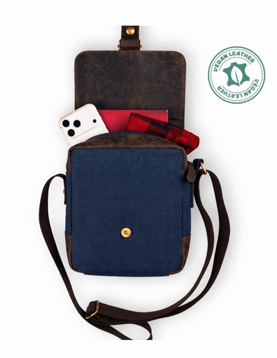 A top-down view of a crossbody phone bag with a navy blue canvas front pocket, dark brown leather trim, and a dark brown leather flap. The bag is shown open with a white smartphone, a red notebook, and a red plaid shirt peeking out.