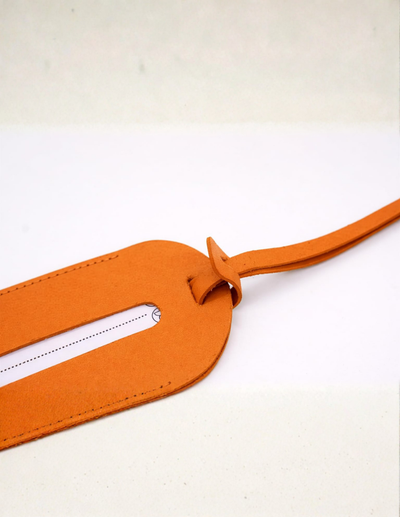 A close-up of an orange leather luggage tag with a long strap, shown on a light off-white background.