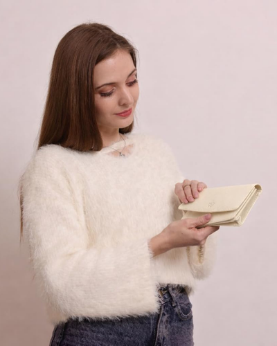 A woman with long brown hair wearing a fluffy white sweater and dark jeans, holding a beige colored leather clutch purse against a solid pale pink background.