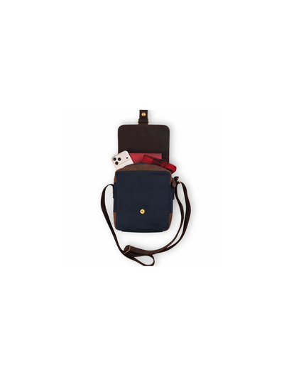 A navy blue canvas crossbody bag with dark brown leather trim, shown open with its strap laid out on a plain white background. A red patterned fabric and a white phone are partially visible inside the compartment.
