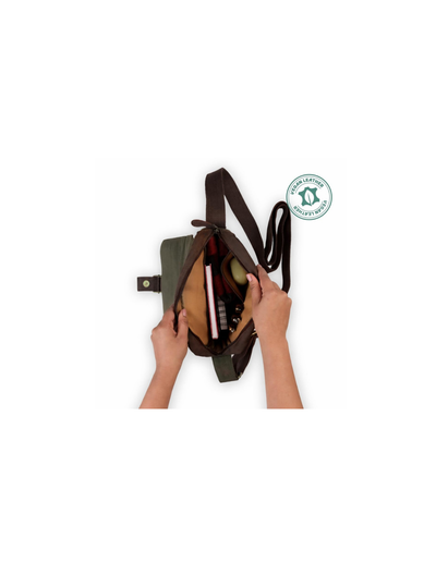 An olive green canvas shoulder bag with a brown interior, shown open with a person's hands holding it. A green 'Fair Trade Certified' logo is displayed on the right side against a solid white background.