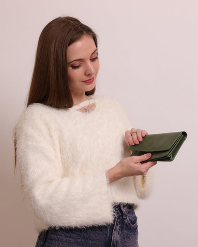 A woman with long brown hair is wearing a fluffy white knit sweater and holding a dark green leather clutch wallet, set against a plain light beige background.