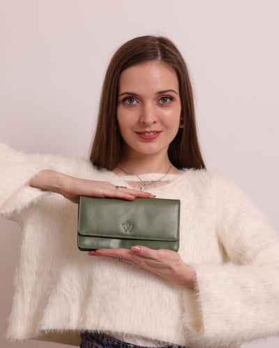 A woman with brown hair wearing a fluffy white sweater holds a green leather clutch bag with a small metallic emblem on the front, set against a solid pale pink background.