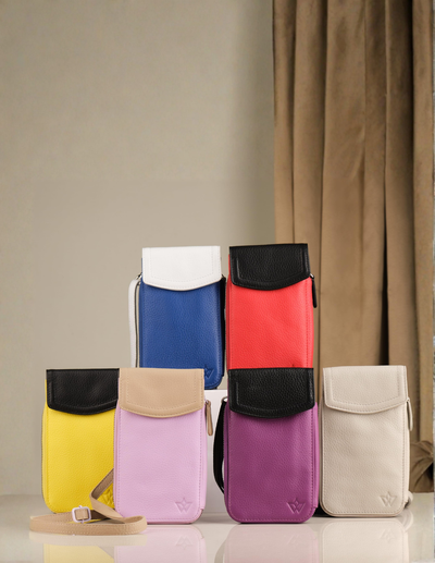Six small flap pouches or phone cases in different colors are arranged on a glossy white surface against a beige wall with brown curtains. The pouches include yellow, light pink, purple, blue, coral red, and off-white, each with a contrasting flap and a small embossed logo near the bottom.