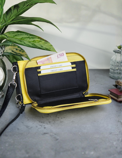 A yellow and black zip-around wallet with a wrist strap, shown open to display its interior card slots and cash compartment, set on a gray tabletop with a plant and small decorative objects in the background.