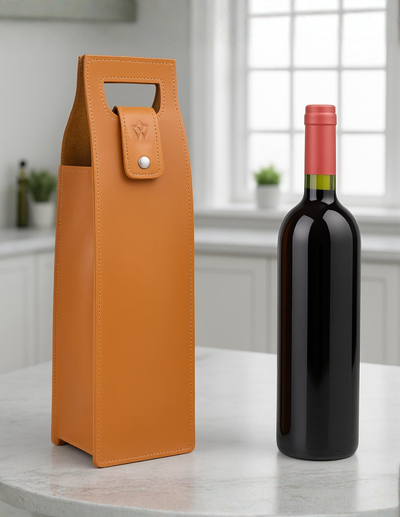 An orange leather wine bottle carrier standing on a light marble countertop next to a dark glass wine bottle with a pink capsule, set against a softly blurred kitchen background.