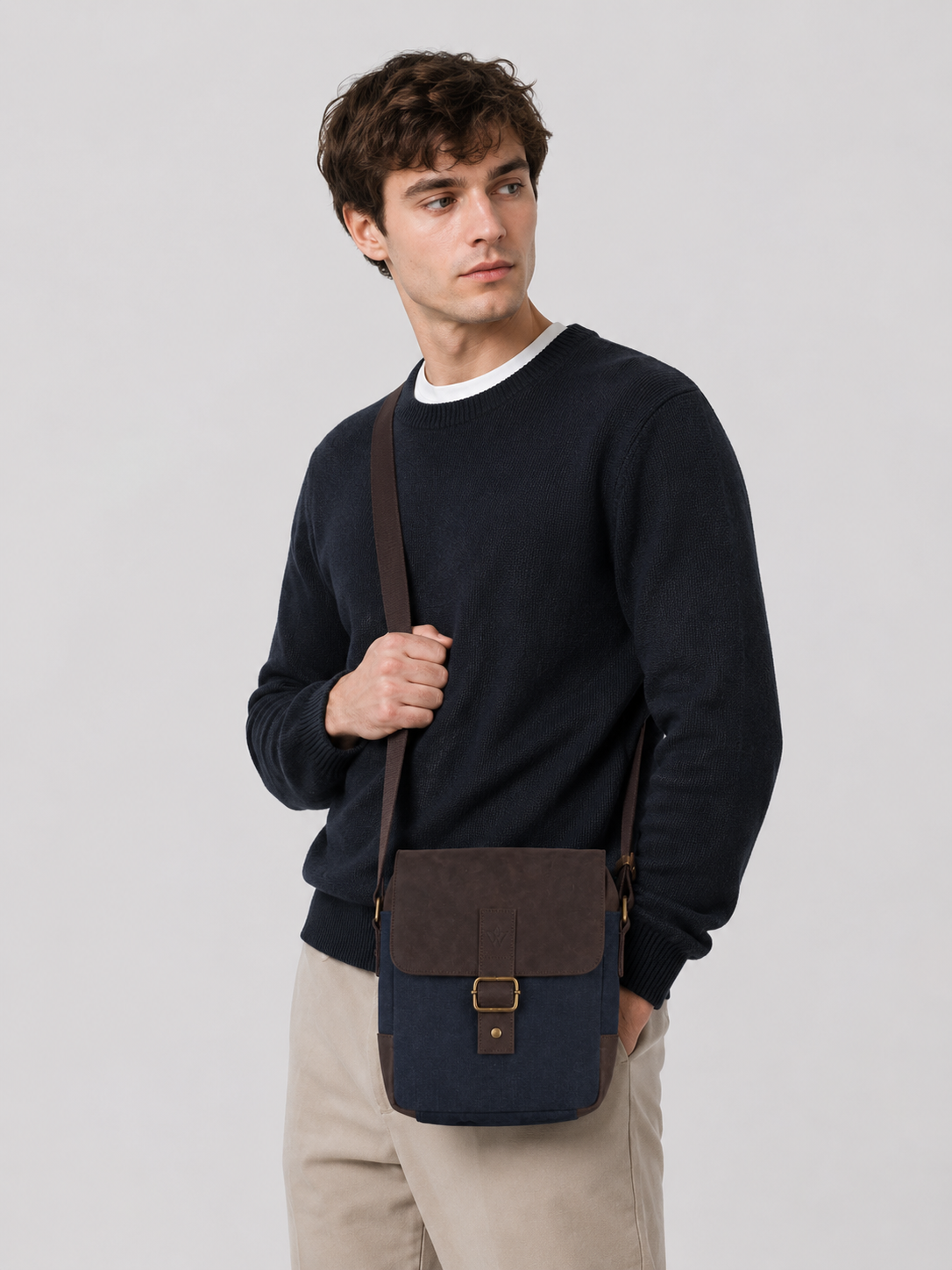 A man wearing a navy blue crewneck sweater and beige trousers carries a dark brown and navy blue crossbody messenger bag with a brown strap and gold-tone buckle against a light gray studio background.