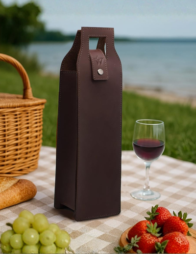 A dark brown leather wine bottle carrier stands on a checkered picnic blanket outdoors, with a glass of red wine, strawberries, grapes, bread, and a wicker basket nearby. A blurred lake and grassy background complete the scene.