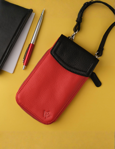 A red and black pebbled leather phone pouch with a black strap, shown on a yellow background next to a black notebook, white paper, and a red and silver pen.