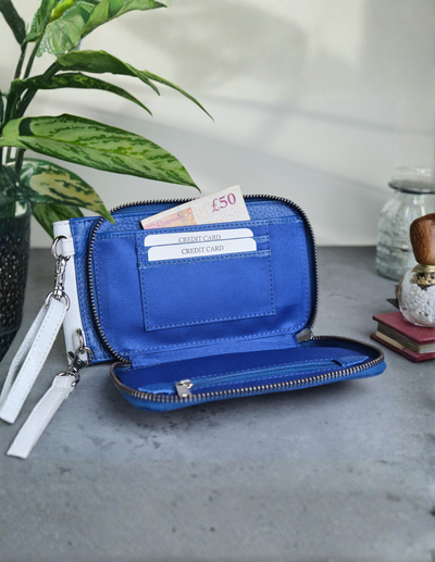 A bright blue zip-around wallet with a wrist strap, shown open on a gray tabletop with a plant and decorative objects in the background.
