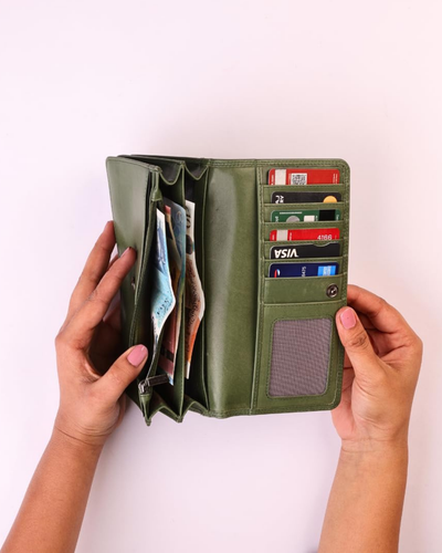 A olive-green colored leather wallet held open by two hands against a light pink background. The wallet is shown with its interior compartments filled with cards and cash, featuring card slots, a gray ID window, and a snap-button closure.