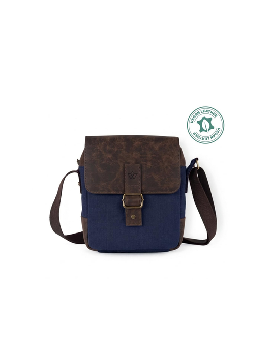 A navy blue canvas crossbody bag with a dark brown textured leather front flap and strap, featuring a brass buckle closure. A green leather certification badge is shown in the upper right corner against a plain white background.